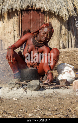 Jeune femme Himba, assis à une fosse de cuisson en face d'une hutte, Kaokoland, Kunene, Namibie Banque D'Images