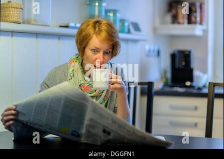 Femme assise à la table de la cuisine à lire le journal, avec un regard étonné, Allemagne Banque D'Images