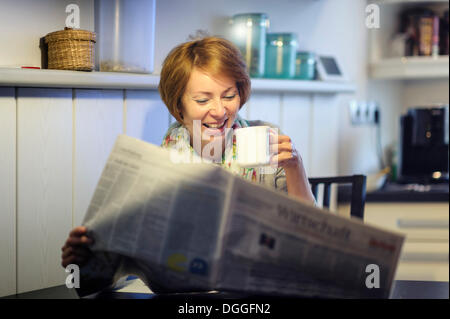 Femme assise à la table de la cuisine à lire le journal, rire, Allemagne Banque D'Images