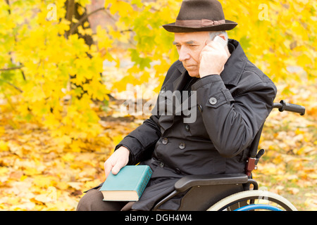 Des personnes âgées handicapées homme assis dans son fauteuil roulant à l'extérieur dans une forêt d'automne jaune coloré avec son téléphone mobile reposant contre sa joue qu'il juge plus de sa récente conversation. Banque D'Images