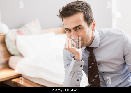 Mid adult man wearing chemise et cravate sitting on bed with hand on chin