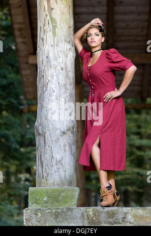 Jeune femme portant un bourgogne dirndl dress posing contre le post en bois d'une cabane forestière, dirndl rechercher Banque D'Images