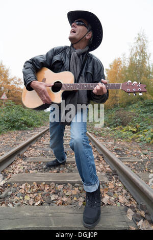 Un homme âgé portant des jeans et une veste en cuir noire avec un chapeau de cow-boy noir et une guitare chant sur une voie ferrée Banque D'Images