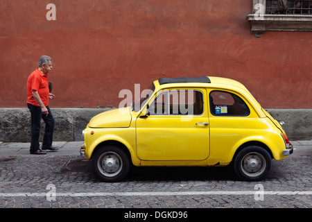 Un homme passe une Fiat 500 jaune vintage voiture garée dans une rue pavée en face d'un mur ocre à Bologne. Banque D'Images