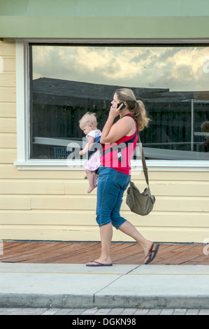Jeune femme mère portant chemise rouge et de pantalons capri carrying baby bébé tout en parlant sur un téléphone cellulaire. Banque D'Images