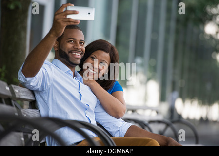 L'été. Un couple assis sur un banc, prenant une photographie selfy. Banque D'Images