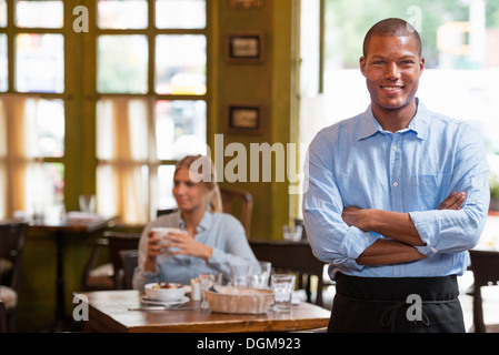 Un couple dans un café de la ville. Une femme s'asseyant la vérification d'un téléphone intelligent. Un homme debout avec les bras croisés. Banque D'Images