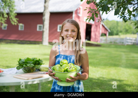 Partie de la famille. Une jeune fille aux longs cheveux blonds portant une robe d'été bleu, portant un grand bol de pommes à peau verte croquante. Banque D'Images