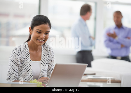 La vie de bureau. Une femme assise à un bureau à l'aide d'un ordinateur portable. Deux hommes dans l'arrière-plan. Banque D'Images