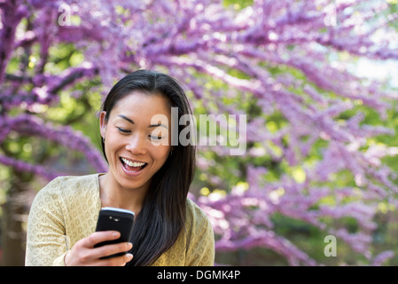 Une femme assise dans un parc à la recherche de son téléphone intelligent et rire. Banque D'Images