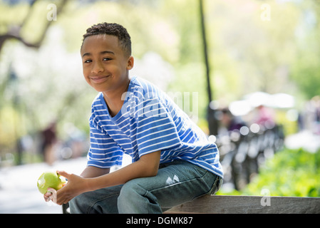 Une famille dans le parc sur une journée ensoleillée. Un garçon assis sur une barrière de manger une pomme. Banque D'Images