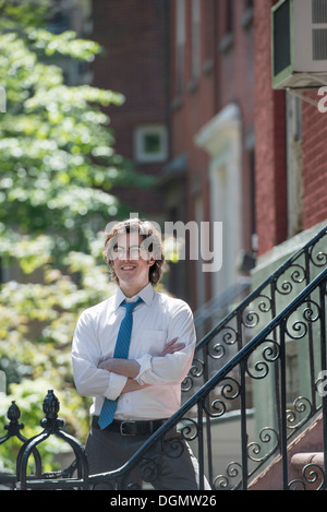 Ville. Un jeune homme en chemise blanche et cravate bleue, debout avec les bras croisés à l'extérieur d'une maison de ville, sur les étapes. Banque D'Images