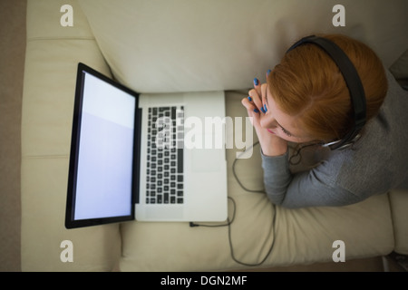 Redhead couché sur la table à l'aide de son ordinateur portable portant des écouteurs Banque D'Images