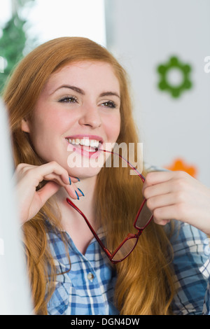 Belle rousse assise à son bureau et de mordre ses lunettes Banque D'Images