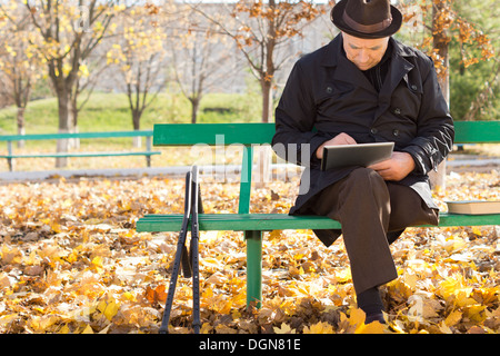 Vieil homme sur des béquilles à l'aide d'un ordinateur tablette comme il est assis dehors au soleil dans un cadre chaleureux sur un manteau d'automne frisquet jour Banque D'Images