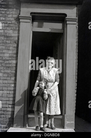 Tableau historique, 1950 d'une grand-mère avec un tablier, tenant les mains de sa petite fille à l'extérieur à la porte d'une maison. Banque D'Images