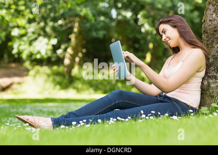 Happy woman leaning against a tree travaillant avec son tablet Banque D'Images