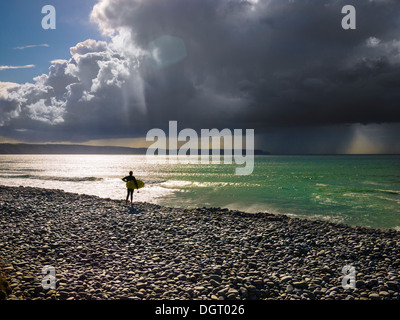 Surfeur se tenait sur une plage de galets avec vue sur la mer avec un orage au loin. Cornborough Range, Abbotsham, North Devon, Angleterre. Banque D'Images