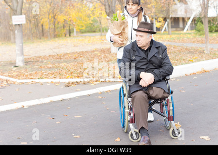 La haute mobilité homme être aidé avec son shopping assis dans son fauteuil roulant comme un soignant ou femelle fille pousse le long de la Banque D'Images