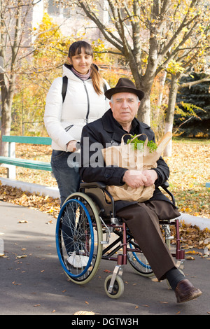 Hauts homme handicapé se rendre à l'épicerie avec l'aide d'une jolie femme souriante qui pousse son fauteuil roulant le long de la rue comme il détient le paquet d'épicerie. Banque D'Images