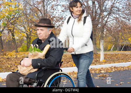 Female carer poussant un vieil homme handicapé dans un fauteuil roulant le long de la rue comme ils reviennent de faire son épicerie. Banque D'Images