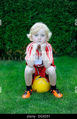 Petit garçon de cinq ans portant une robe de football FC Bayern Munich, assise sur un ballon jaune Banque D'Images