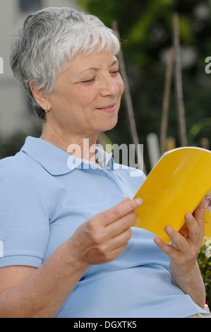 Une femme âgée, aux cheveux gris, 55-65, lire un livre à l'extérieur Banque D'Images