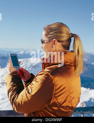 Femme portant des vêtements d'hiver tenue d'un téléphone cellulaire sur la montagne enneigée, pics, Zugspitze Wetterstein, Alpes orientales Banque D'Images