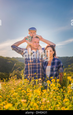 Une jeune famille caucasienne, père, mère et bébé garçon sur les épaules de papa, se promenant dans un champ de fleurs jaunes. Banque D'Images