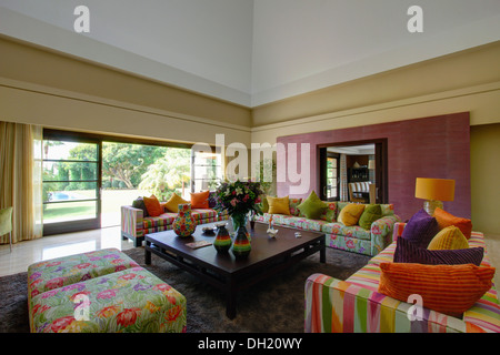 Des coussins colorés et fleuris sur canapés et tabourets de bar rayé dans le salon moderne avec une table basse en verre et des portes Banque D'Images