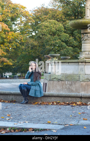 Jeune femme dans un parc en automne assise sur le bord d'une fontaine à l'aide d'un iphone d'Apple pour effectuer un appel Banque D'Images