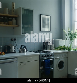 Avant de l'armoire en verre opaque sur le mur au-dessus de cuisinière à gaz et un petit lave-linge en bleu pâle cuisine avec bouilloire et grille-pain chromé Banque D'Images