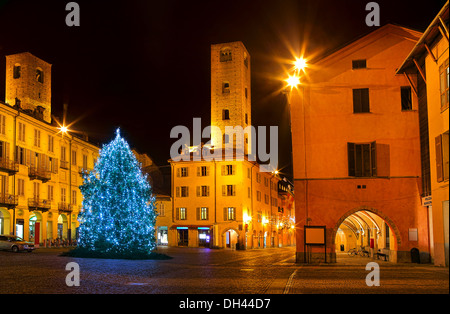 Arbre de Noël décoré et illuminé sur la place centrale de la ville entourée de maisons anciennes et les tours médiévales de nuit à Alba. Banque D'Images