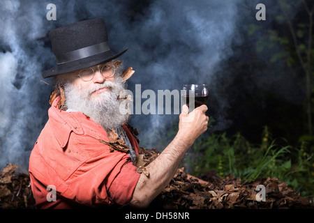 Un homme âgé avec une longue barbe et vêtu d'un haut chapeau et lunettes, couché dans un tas de feuilles d'automne et tenant un verre de vin rouge Banque D'Images