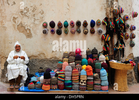 Vendeur de rue portant une Djellabah traditionnelle tout en tricotant une PAC, à côté de lui sont ses chapeaux pour vente, Essaouira, Maroc Banque D'Images