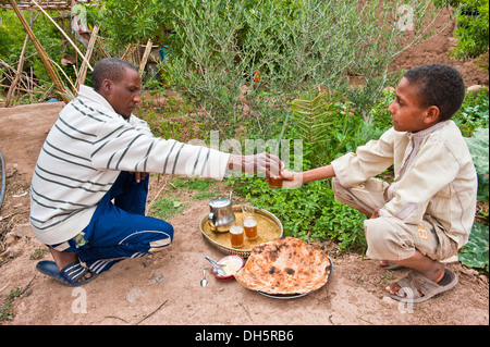 Un homme Berbère est de remettre un verre de thé à la menthe à un garçon, sur le terrain il y a télévision pain, beurre et un plateau avec une théière et laiton Banque D'Images