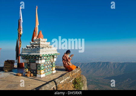 Sadhu ou saint homme assis avec ses mains jointes en prière sur le précipice devant un temple hindou de culte avec les drapeaux de prières Banque D'Images