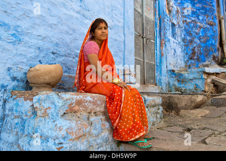 Femme âgée portant un sari rouge assis sur une corniche d'un bâtiment résidentiel peinte en bleu, Brahmpuri, Jodhpur, Rajasthan, India Banque D'Images