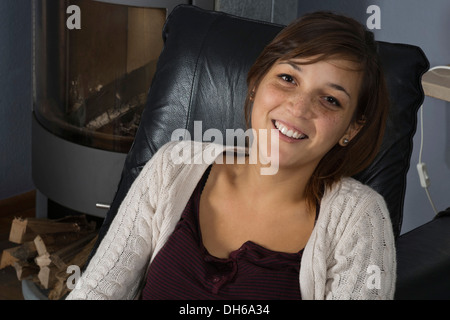 Une jeune femme d'origine indonésienne est assis sur un fauteuil en cuir en face d'un poêle à bois, de détente Banque D'Images