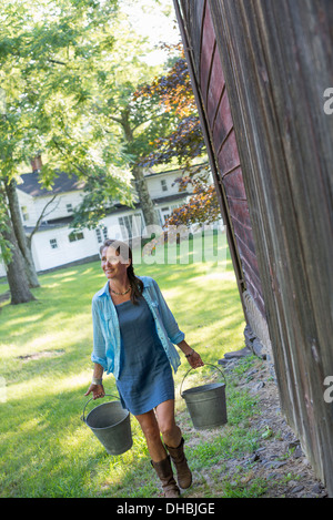Une femme portant un seau pour l'alimentation animale dans chaque main, à l'extérieur une grange. Banque D'Images