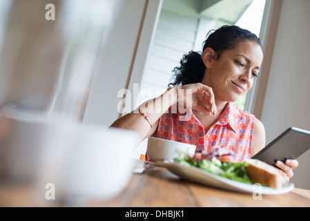 Une jeune femme en train de lire l'écran d'une tablette numérique, assis à une table. Le café et un sandwich. Banque D'Images