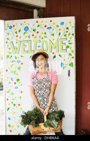 Travailler sur une ferme biologique. Une jeune femme asiatique par le panneau de bienvenue avec un grand panier de légumes, fraîchement cueillies. Banque D'Images