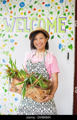 Travailler sur une ferme biologique. Une jeune femme asiatique par le panneau de bienvenue avec un grand panier de légumes, fraîchement cueillies. Banque D'Images