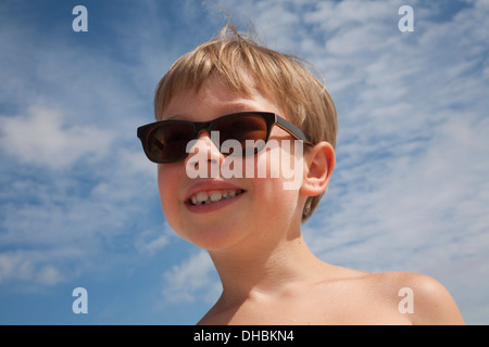 Un jeune garçon portant des lunettes de soleil. Smiling, ciel bleu. Les cheveux blonds. Banque D'Images