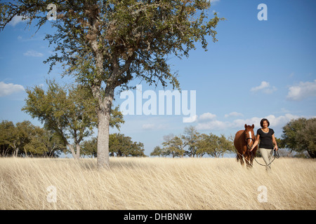 Une femme à la tête d'un cheval sur le dos à travers les hautes herbes dans un champ. Banque D'Images