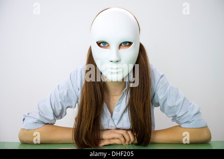 Fille brune aux yeux bleus avec des yeux effrayés portant un masque blanc avec un sourire créepy photographié sur un fond blanc mains sur la table Banque D'Images