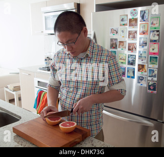 Un homme asiatique tranches un pamplemousse rose en face d'un réfrigérateur en acier plein de photos Polaroid dans un appartement lumineux, cuisine. Banque D'Images