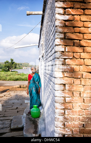 Personnes âgées femme indienne portant un pot en plastique avec de l'eau d'une borne fontaine dans son village house. L'Andhra Pradesh, Inde Banque D'Images