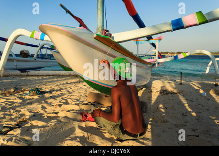 Garçon indonésien assis sur une plage de sable sur la conception de peinture proue d'un outrigger voilier Banque D'Images