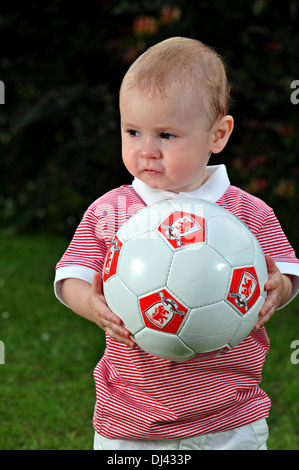 Un jeune garçon, âgés de deux, est titulaire d'un ballon de soccer qu'il en a l'air de l'appareil photo et sourit légèrement. Banque D'Images